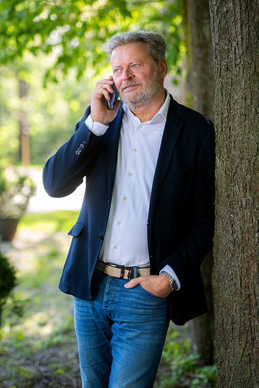 Makelaar staat buiten met zijn telefoon langs zijn oor en leunend tegen een boom