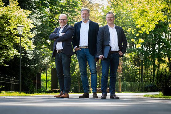 Drie makelaars lachend op de foto, staan midden op een lange weg met veel bomen