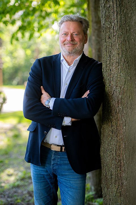 Makelaar staat glimlachend geleund tegen een boom